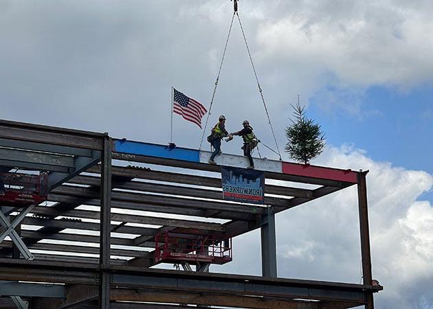 2 people stand on a red, white, and blue beam on one corner of a steel framed structure. they are shaking hands. an evergreen tree is on one end and a United States flag is on the other.