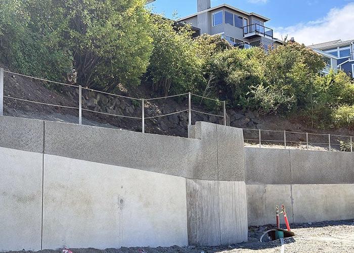 a two-level concrete retaining wall has a railing on top and there is shrubbery above and houses visible above that