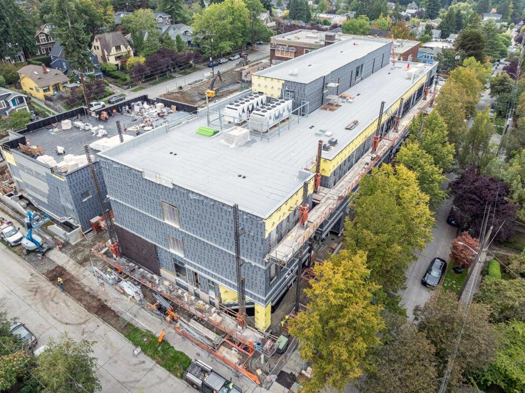 a large 3 story building under construction with gray material on the outside and trees next to it with green leaves